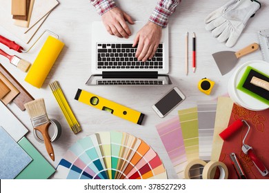 Professional Decorator's Hands Working At His Desk And Typing On A Laptop, Color Swatches, Paint Rollers And Tools On Work Table, Top View