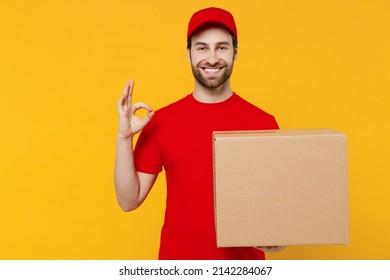Professional delivery guy employee man 20s in red cap T-shirt uniform workwear work as dealer courier hold face with cardboard box show ok gesture isolated on plain yellow background. Service concept