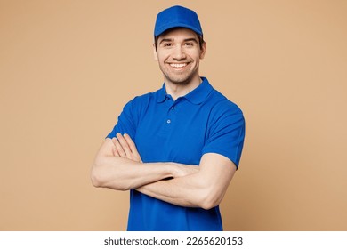 Un empleado profesional de reparto lleva puesto ropa de trabajo uniforme de camiseta de gorro azul mientras la cámara de aspecto de mensajero de la repartidor sujeta las manos cruzadas plegadas aisladas sobre fondo beige claro. Concepto de servicio