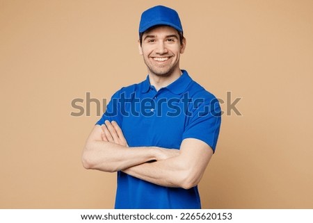 Professional delivery guy employee man wears blue cap t-shirt uniform workwear work as dealer courier look camera hold hands crossed folded isolated on plain light beige background. Service concept
