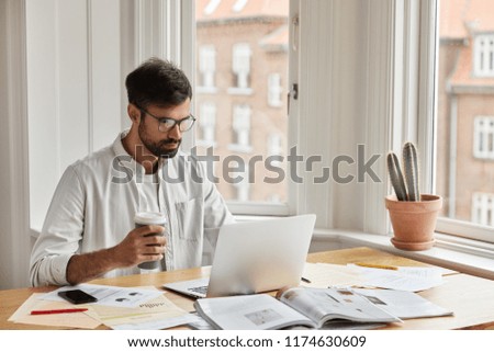 Professional IT developer works on design of new site, studies documentation, holds takeaway coffee. Focused concentrated young graphic designer develops new strategy, poses in cozy interior