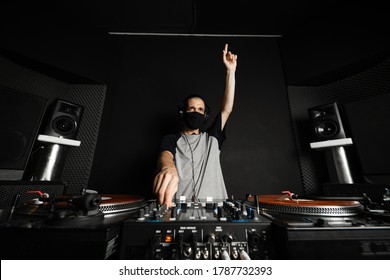 Professional DJ with reusable cloth face mask playing music raising his hand to cheer up the clients of the disco, with orange records and a gray T-shirt with dark blue sleeves