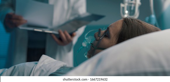 Professional Doctor Assisting A Patient Lying In Bed At The Hospital With An Oxygen Mask, He Is Checking Medical Records