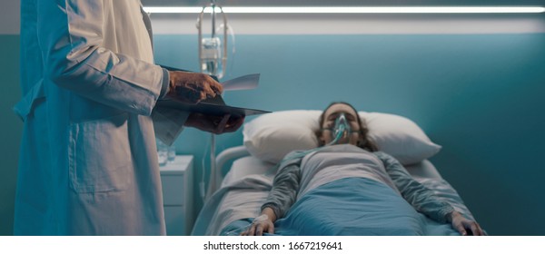 Professional Doctor Assisting A Patient Lying In Bed At The Hospital With An Oxygen Mask, He Is Checking Medical Records