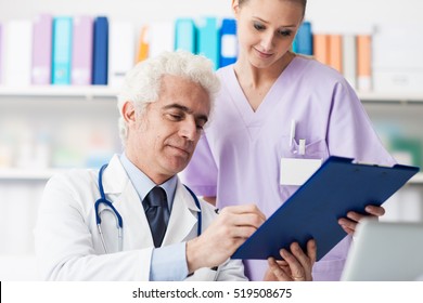 Professional Doctor And His Assistant Working In The Office, She Is Holding A Clipboard And He Is Signing Medical Records