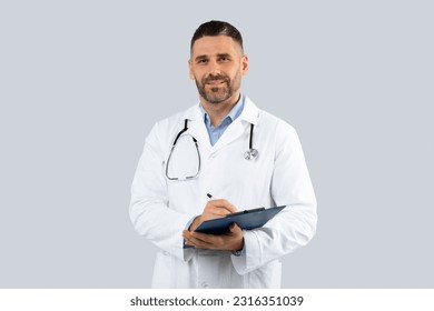 Professional doctor man in medical coat with clipboard in hands posing over light grey wall background, middle aged physician with stethoscope looking at camera - Powered by Shutterstock