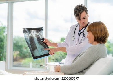 Professional Doctor In The White Coat Put A Stethoscope Around Neck Holding X-ray Film And Talked To Elderly Woman Who Lying Down On The Bed At Nursing Home. Explanation On The Film Result To Patient