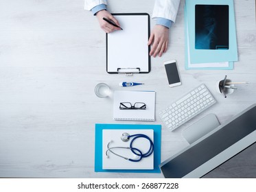 Professional Doctor Writing Medical Records On A Clipboard With Computer And Medical Equipment All Around, Desktop Top View With Copyspace