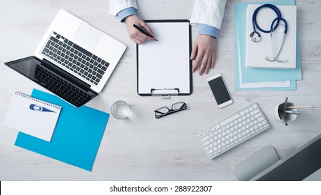 Professional Doctor Writing Medical Records On A Clipboard With Computer And Medical Equipment All Around, Desktop Top View