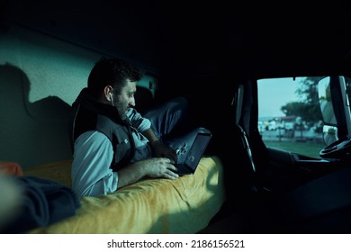 Professional Driver Relaxing In Truck Cabin While Surfing The Net On Laptop At Night On Parking Lot. 