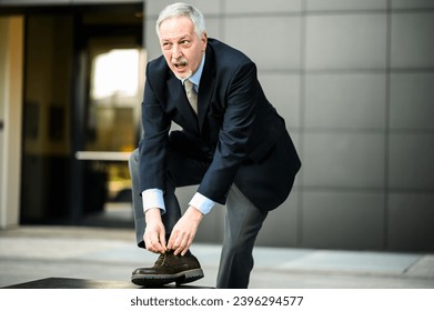 Professional elderly man in a suit bending over to tie his shoe outdoors - Powered by Shutterstock