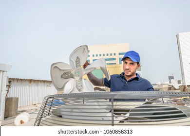 A Professional Electrician Man Is Showing The Broken Fan Of A Heavy Duty Unit Of An Air Conditioner And Wearing Blue Uniform And Cap At The Roof Top And City Buildings In The Background 