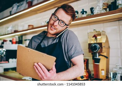 Professional employee of cafeteria dressed in black apron calling on smartphone device and taking order from client during phone conversation standing at bar.Barista talking on modern cellular - Powered by Shutterstock