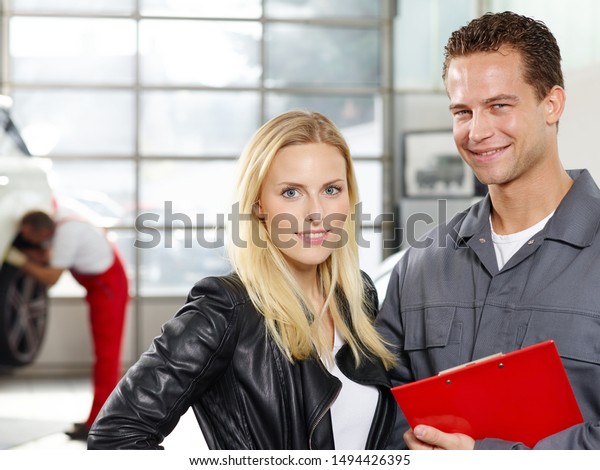 a\
professional employee of a tire and car service workshop stand with\
a clipboard and a nice smile next to the blonde female customer and\
you see the mechanical work in the\
background