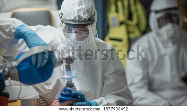 Professional EMS Paramedic in Disposable\
Coverall Suit Provide Medical Help to Injured Patient on the Way to\
Hospital. Emergency Care Assistant Putting Non-Invasive Ventilation\
Mask in an\
Ambulance.