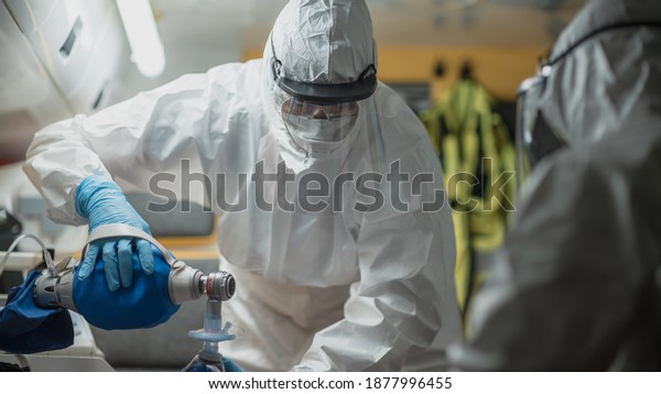 Professional EMS Paramedic in Disposable\
Coverall Suit Provide Medical Help to Injured Patient on the Way to\
Hospital. Emergency Care Assistant Putting Non-Invasive Ventilation\
Mask in an\
Ambulance.