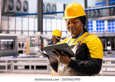 Professional engineer with tablet, enhancing efficiency of automated machinery in industrial production plant. Black female industry 4.0 expert overseeing manufacture of robot arms in smart factory.
