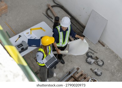 Professional engineer and worker stand in construction site holding blueprint wearing hardhat inside an uncompleted building displaying focused teamwork on development project