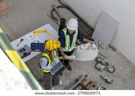 Professional engineer and worker stand in construction site holding blueprint wearing hardhat inside an uncompleted building displaying focused teamwork on development project