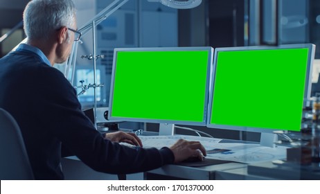 Professional Engineer Works On Computer With Two Green Mock-up Screen Displays. In The Background Industrial Electronics Design Agency. Over The Shoulder Shot