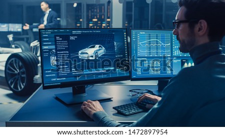 Professional Engineer Works on a Computer with a 3D CAD Software and Tests the Electric Car Chassis Prototype with Wheels, Batteries and Engine Standing in a High Tech Development Laboratory.