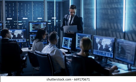 Professional IT Engineers Working In System Control Center Full Of Monitors And Servers. Supervisor Holds Laptop And Holds A Briefing. Possibly Government Agency Conducts Investigation. 