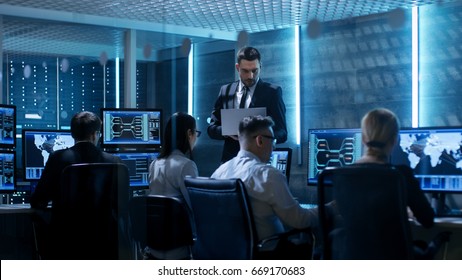 Professional IT Engineers Working in System Control Center Full of Monitors and Servers. Supervisor Holds Laptop and Holds a Briefing. Possibly Government Agency Conducts Investigation. 