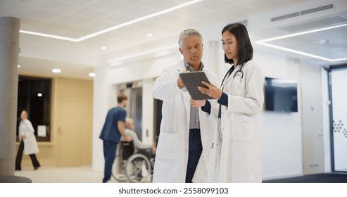 Professional Exchange Between a Caucasian Medical Laboratory Scientist and a Young Multiethnic Female Doctor in a Hospital Hall. They Review Patient Care Data on a Tablet Computer Together as a Team - Powered by Shutterstock