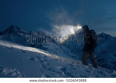 Professional expeditor commit climb on snowy mountains at night and lights the way with a headlamp. Wearing ski wear, backpack and a snowboard behind his back. Backcountry and ski touring