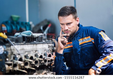 Professional experienced car mechanic repairman in uniform thinking about solution and looking at car engine in mechanics workshop. Repairing car.