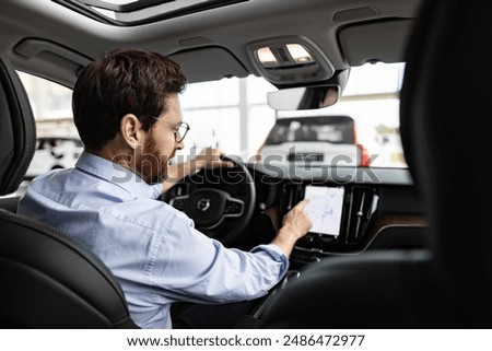 Professional explores car features preparing for purchase in bright showroom. Businessman interacts with car touchscreen sitting inside vehicle in dealership.