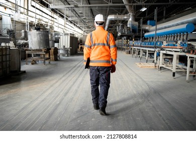 Professional factory worker or engineer in high visibility jacket and hardhat walking through industrial production hall with machinery. Factory interior. - Shutterstock ID 2127808814