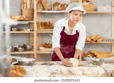 Professional female baker in maroon apron kneading dough on floury table in artisan bakery shop, surrounded by freshly baked bread and pastries arranged on wooden shelves - Powered by Shutterstock