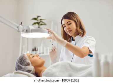 Professional female beautician makes a rejuvenating injection to a young woman who lies relaxed with her eyes closed in a modern beauty salon. Concept of smoothing wrinkles with injections.