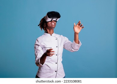 Professional female chef in uniform playing virtual reality games with VR goggles, exploring simulations. African american cook moving arms while touching objects in cyberspace. - Powered by Shutterstock