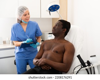 Professional Female Cosmetologist Planning Upcoming Treatments For African American Male Patient In Modern Aesthetic Medicine Office