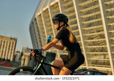 Professional Female Cyclist In Black Cycling Garment And Protective Gear Holding Water Bottle While Riding Bicycle In City, Training Outdoors On A Warm Day