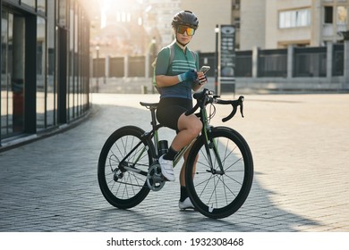 Professional female cyclist in cycling garment and protective gear looking at camera, holding smartphone while riding bicycle in city center. Sportswoman training, exercising outdoors - Powered by Shutterstock