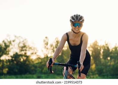 Professional female cyclist in cycling garment and protective gear riding bicycle in the nature during a sunset. Training outdoors on a daytime for a race. Smiling while looking into the camera. Flare - Shutterstock ID 2199514373