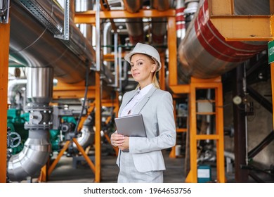 A professional female director in formal attire, with a protective helmet on her head holding a tablet and standing in a heating plant. She stands at the entrance of the heating plant. CEO female - Shutterstock ID 1964653756