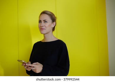 Professional Female Employee Thinking About Something Good While Standing With Mobile Phone In Office Interior, Young Businesswoman Holding Cell Telephone In Hands And Looking Away During Work Break 