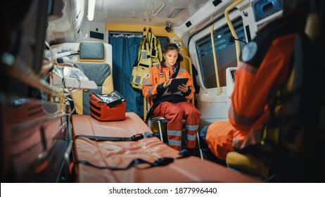 Professional Female EMS Paramedic Using Tablet Computer While Riding In An Ambulance Vehicle For An Emergency. Emergency Medical Technicians Are On Their Way To A Call Outside The Healthcare Hospital.