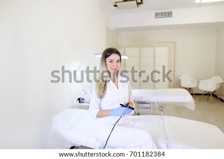 Professional female eyebrow tattooist, attractive young woman holding black mechanical machine with needles in hands to clean eyebrows, looks into camera and smiles, sitting on white couch in modern