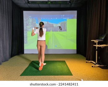 Professional female golfer holding club playing golf indoors on golf simulator. Driving range with virtual golf screen - Shutterstock ID 2241482411