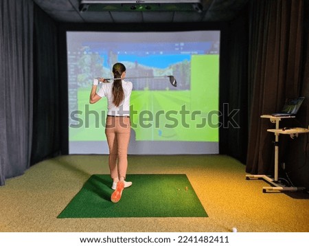 Professional female golfer holding club playing golf indoors on golf simulator. Driving range with virtual golf screen