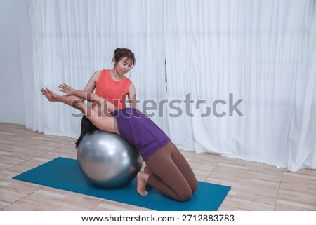 Professional female instructor assisting a client with back stretching exercise using a large Pilates ball on yoga mat.