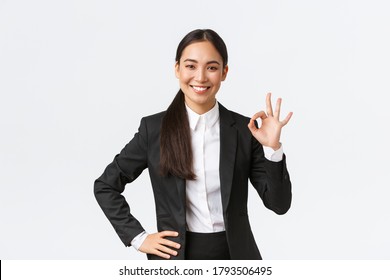 Professional female manager in black suit looking assertive, encourage everything okay, assure work done, showing okay gesture and smiling in approval, standing satisfied over white background