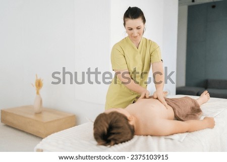 Professional female pediatric masseuse making therapeutic massage on tummy to five year old boy in clinic. Rehabilitation massage on stomach of child lying on massage table. Concept of childcare.