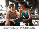 Professional female personal trainer talking with male client, discussing nutrition or training plan on clipboard in modern gym interior