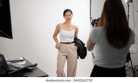 A professional female photographer is taking pictures of an attractive young Asian female model in casual wear, having a fashion photoshoot in a studio with professional lighting equipment. - Powered by Shutterstock